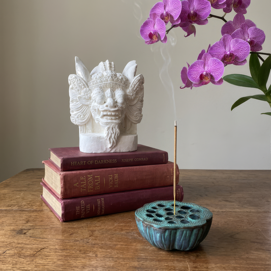Lotus Pod ashtray by TIBA-TIBA – Solid bronze lotus incense burner with Barong sculpture sitting on books and purple orchid in background.