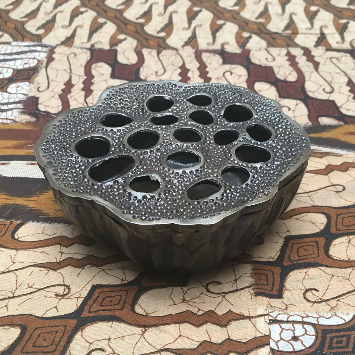 Lotus Pod ashtray by TIBA-TIBA – Close up detail of sculptural bronze ashtray in shape of a lotus seed, used as paperweight or incense burner on brown Batik fabric. 