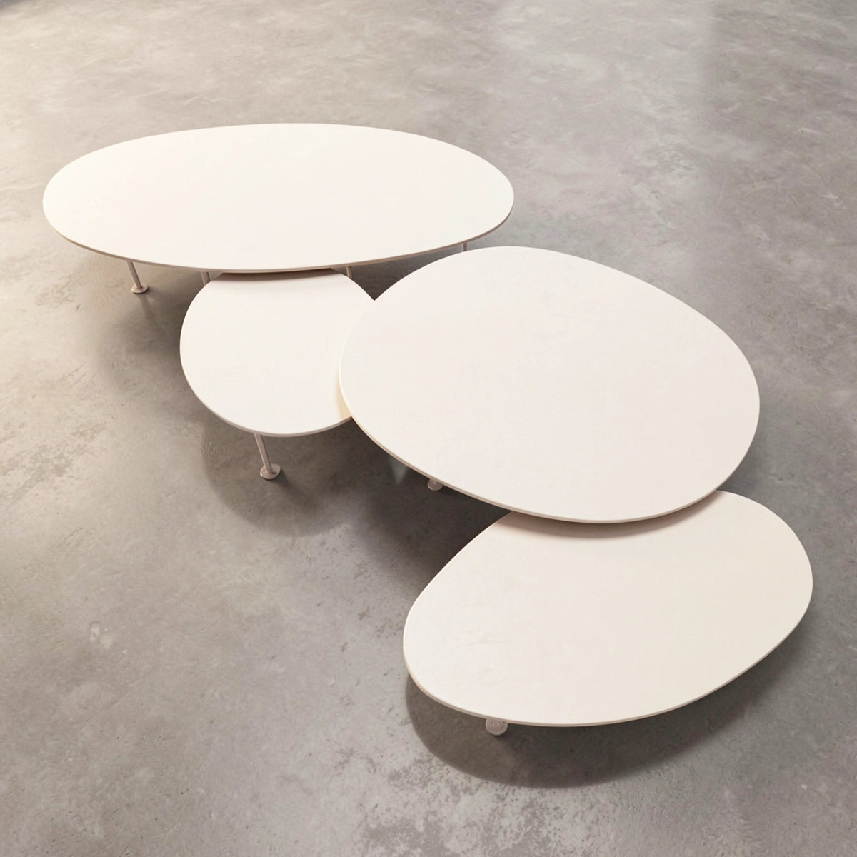 A set of four nested, organic-shaped Pebbles - Coffee Tables by TIBA-TIBA in a warm off-white finish of Dekton Aeris, resting on a textured grey concrete floor.