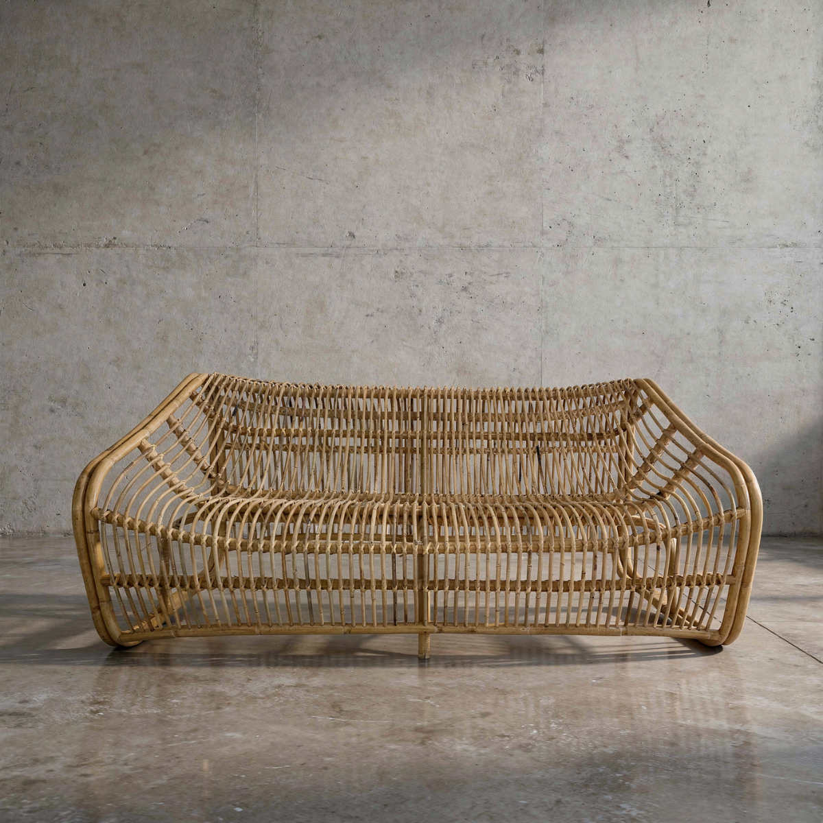 Bucket 2-Seater Sofa by TIBA-TIBA – wicker two-seat lounge sofa with sculptural curved frame on polished concrete floor with concrete wall background. 
