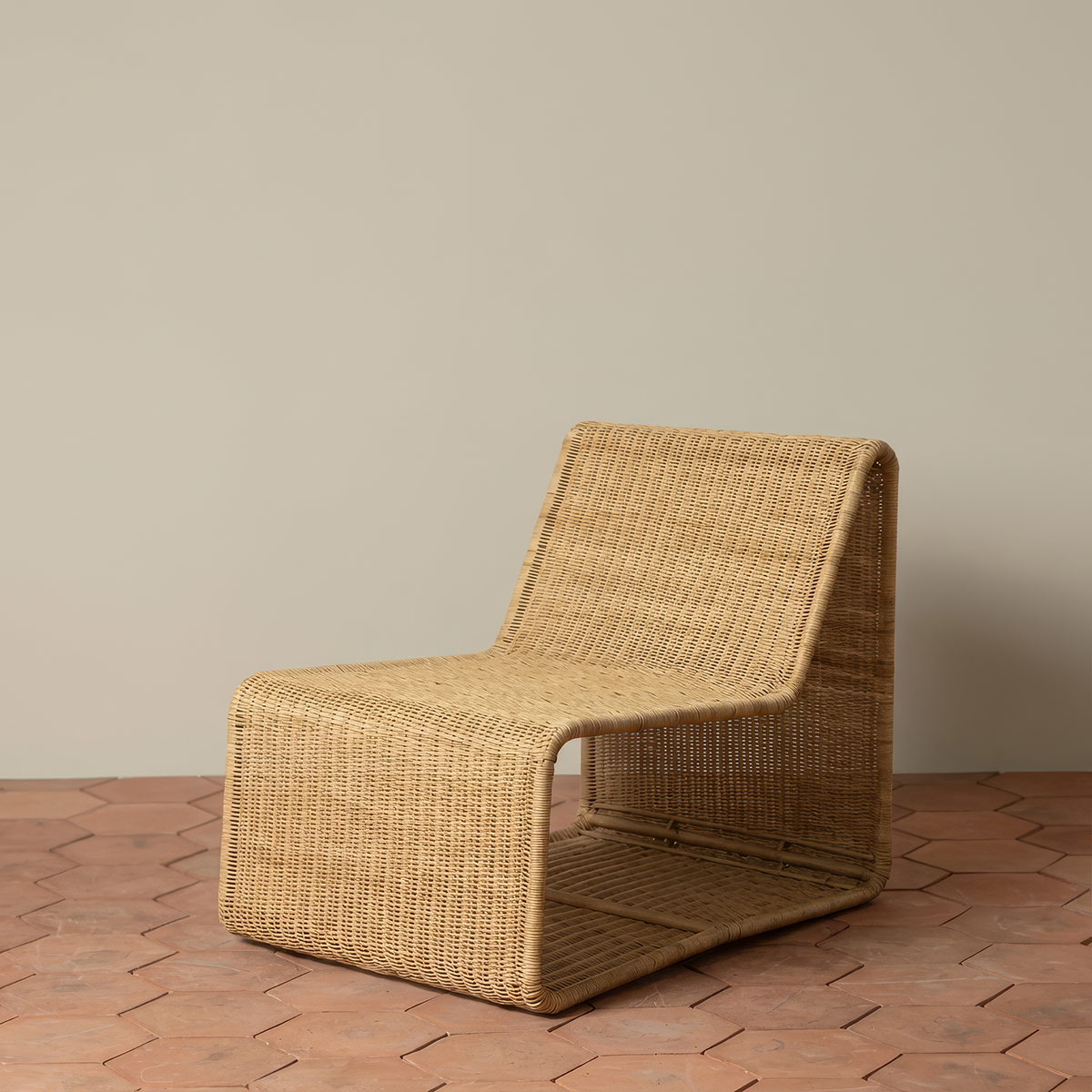 Geo Lounge Chair by TIBA-TIBA – handwoven rattan lounge chair in a geometric cube form with natural honey finish on terracotta red hexagon tiles and taupe background wall. 