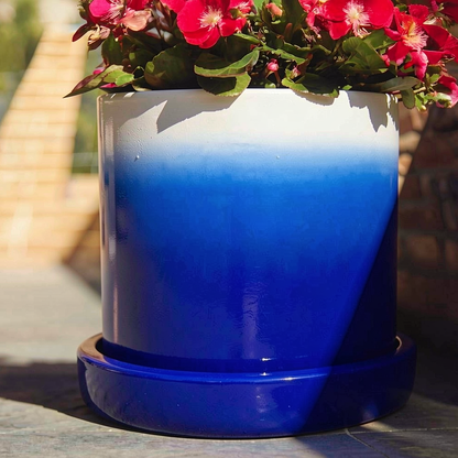 Fade Planter and Tatakan Plant Saucer by TIBA-TIBA - A dark blue ombre planter with a dark blue matching ceramic drip tray on an outdoor table with pink flowers. 